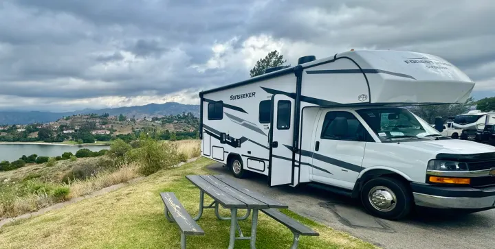 This white Forest River Sunseeker RV rental is parked by a picnic table, overlooking a scenic lake, distant hills, and a cloudy sky.. Forest River Sunseeker 2026