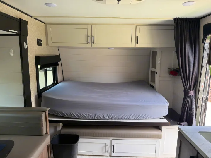 Compact RV rental interior showing a bed with gray sheets, overhead cabinets, window, dinette seating, and storage.. Autumn Ridge 188BHS 2025