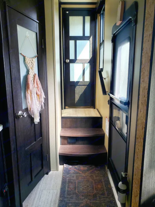 Inside this cozy RV rental, steps lead up to a sunlit main door. Another door with macrame decor and a fire extinguisher are visible.. Grand Design Momentum 2017