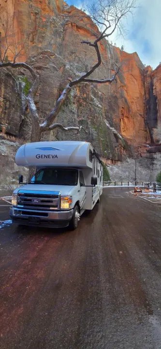 A white **rv rental** is parked on a wet road in front of towering, snow-dusted red rock canyon walls and bare winter trees.. Thor Geneva 2025