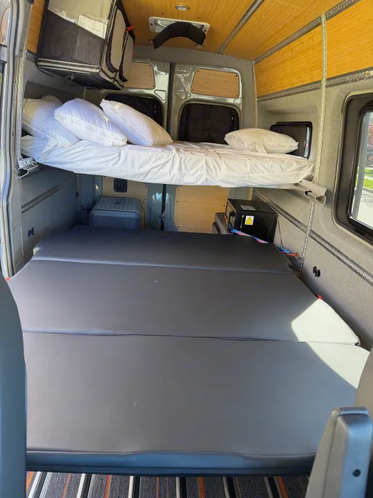 Spacious rv rental interior featuring a raised bed with "Roots Home" pillows and flexible lower cushioned sleeping/seating.. Mercedes-Benz Sprinter 2025