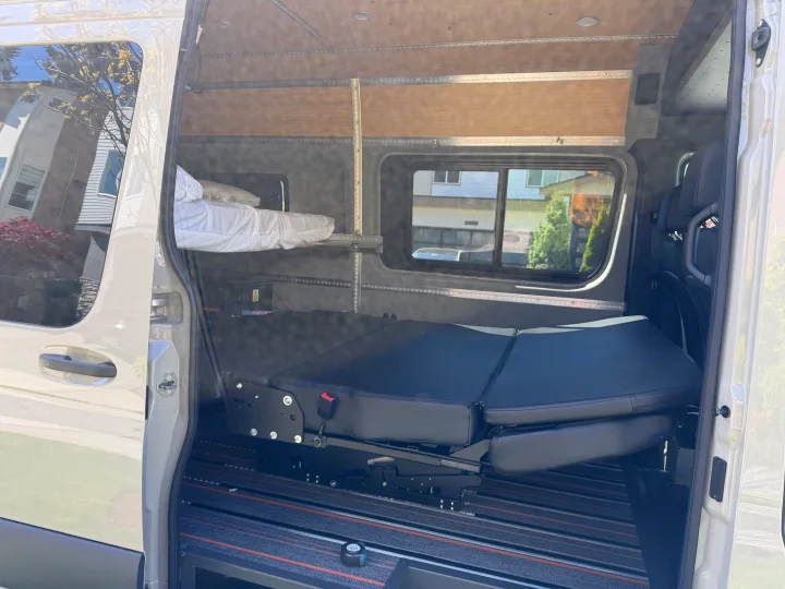 Open side door reveals a well-equipped rv rental interior with a foldable bench/bed, upper bunk, and modular track system for travel.. Mercedes-Benz Sprinter 2025