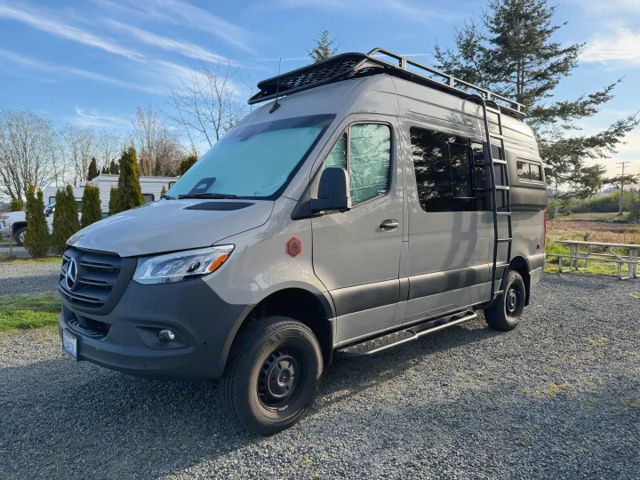 Gray Mercedes Sprinter RV rental van, with a roof rack and ladder, parked on gravel, ready for adventure.. Mercedes-Benz Sprinter 2025
