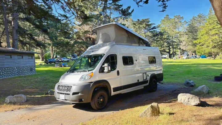 This rv rental, a white camper van with a pop-top, is parked at a sunny forested campground with other campers visible.. Winnebago Solis 2022