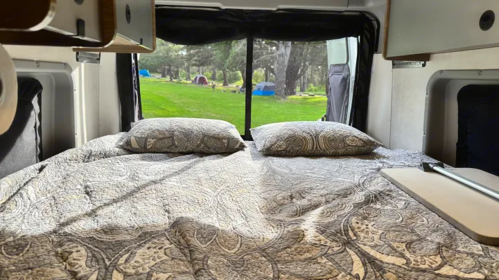 Inside an RV rental, a comfy bed faces a large window overlooking a green campsite with tents and trees outside.. Winnebago Solis 2022