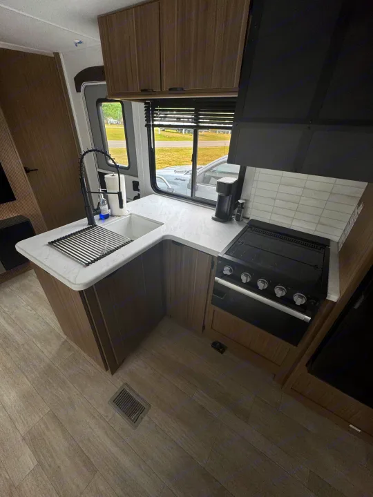 Modern RV rental kitchen featuring a stylish sink with drying rack, stovetop, coffee maker, wood cabinets, and light countertops.. Forest River Wildwood 2025