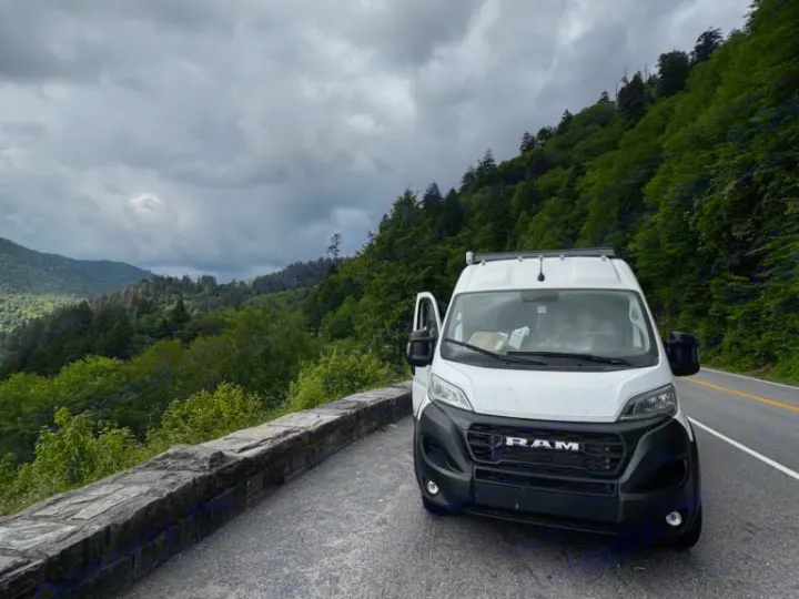 Our white Ram ProMaster RV rental on a mountain road, with cloudy skies and lush forests. Adventure awaits!. Dodge Other 2023