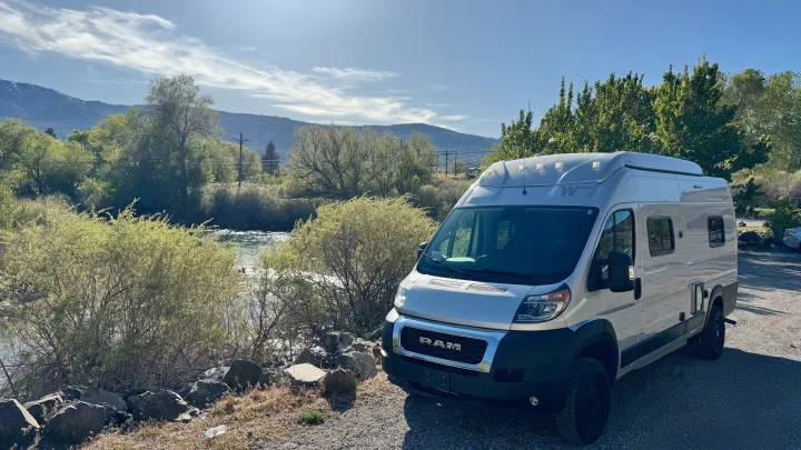 Experience nature with this RV rental! A silver camper van is parked by a serene river with mountains and green trees under a bright sky.. Winnebago Solis 2022