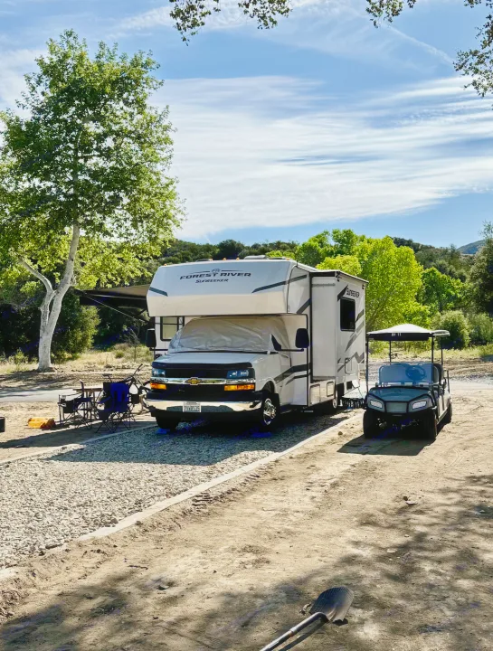 A white Forest River Sunseeker rv rental and a golf cart are parked at a sunny campsite with trees and a blue sky.. Forest River Sunseeker 2026