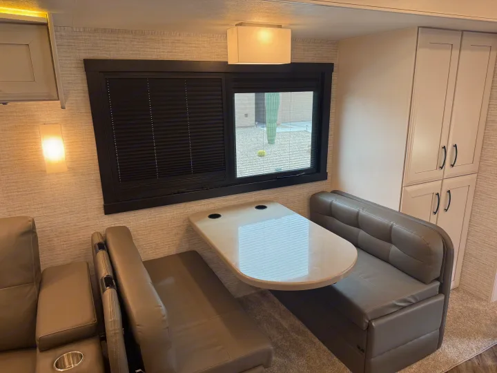 Modern RV rental interior: a comfy dinette with grey benches, a table with cup holders, and a window looking out to desert scenery.. Entegra Coach Accolade 2025