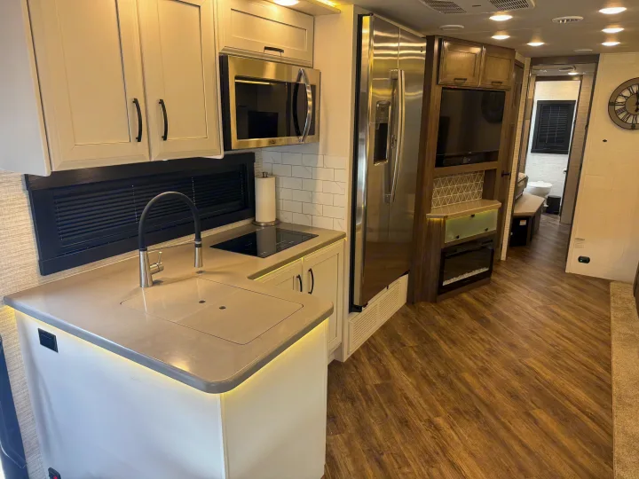 Modern RV rental kitchen: light cabinets, illuminated island, stainless appliances, wood floor. Living area & bath beyond.. Entegra Coach Accolade 2025