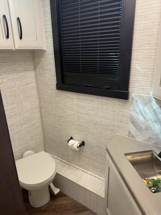 RV rental bathroom features a compact toilet, sink, white cabinets, textured walls, and a window with black blinds.. Entegra Coach Accolade 2025