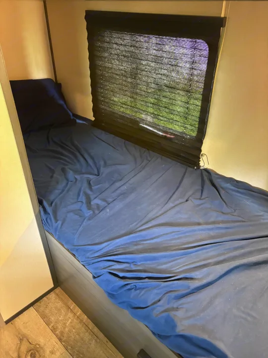 This RV rental bunk features dark blue bedding, a pillow, and a window with a mesh blind overlooking green trees.. Aspen Trail Aspen Trail Trailer 2022