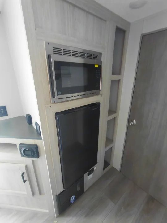 RV rental kitchen featuring a built-in GE microwave, Insignia mini-fridge, storage, and electrical panel in light wood.. Coachmen Catalina Summit Series 2024