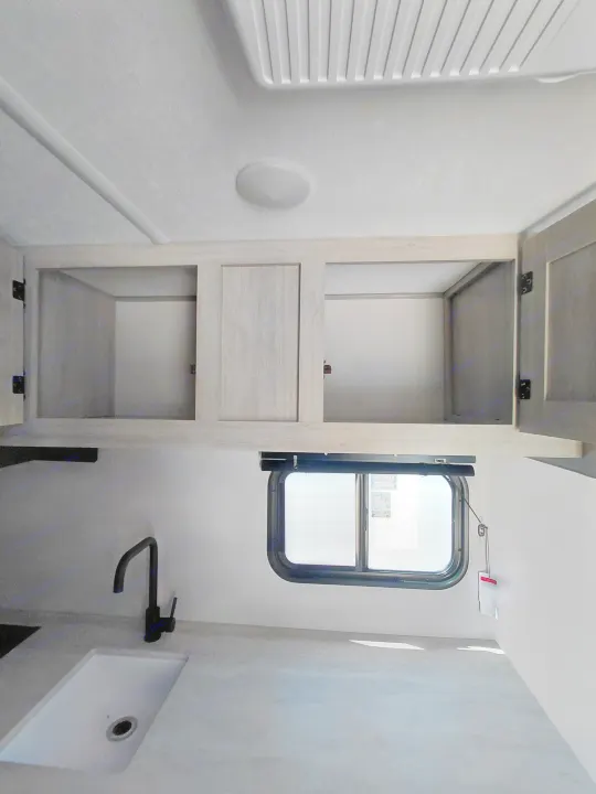 RV rental kitchen view: light wood-grain overhead cabinets, modern black faucet, sink, and a window with a shade. Clean design.. Coachmen Catalina Summit Series 2024