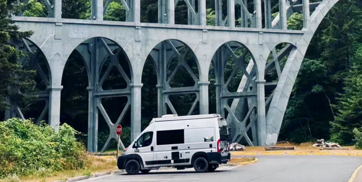 An RV rental camper van parked under a historic arched concrete bridge, surrounded by dense evergreen forests and cloudy skies.. Thor Motor Coach 18T 2022