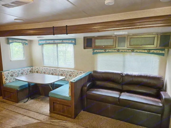 RV rental interior showing a dinette booth, table, and dark leather sofa. Windows with blinds and overhead cabinets are visible.. Heartland RVs Prowler 2018