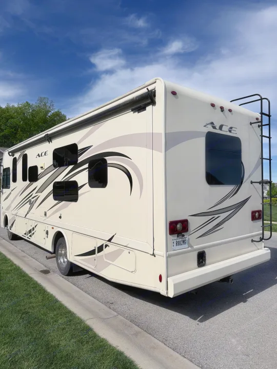 Cream ACE **RV rental** parked curbside. Its side features black/gray graphics, an awning, windows, and a rear ladder under a blue sky.. Thor A.C.E. 2018