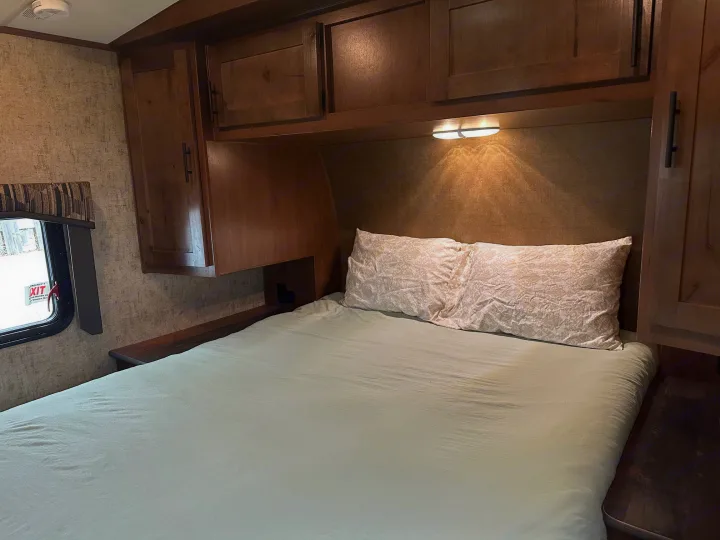 Comfortable RV rental bedroom featuring a bed, wood cabinets, and an overhead reading light. Emergency exit window visible.. Outdoors RV Timber Ridge 2020