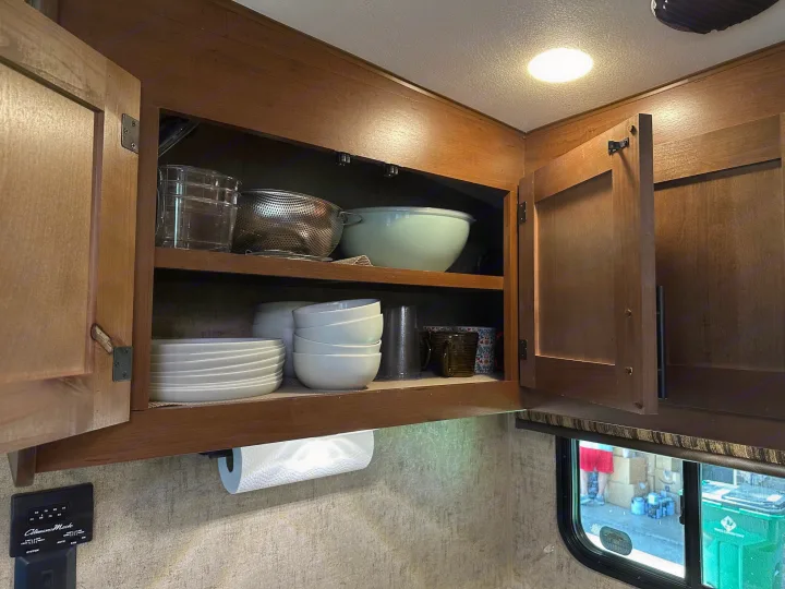 RV rental kitchen: An open cabinet reveals neatly stacked dishes, bowls, and a colander. A paper towel roll hangs below, next to a window.. Outdoors RV Timber Ridge 2020