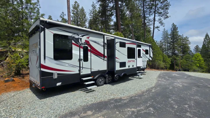 Large gray and red Raptor fifth-wheel RV rental parked on a gravel lot amidst tall pine trees.. Keystone RV Raptor Toy Hauler 2015