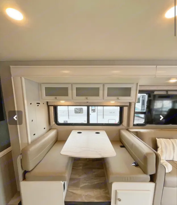 Comfortable RV rental dinette featuring bright lighting, tan bench seats, a light-colored table, and white overhead cabinets.. Holiday Rambler Admiral 2021