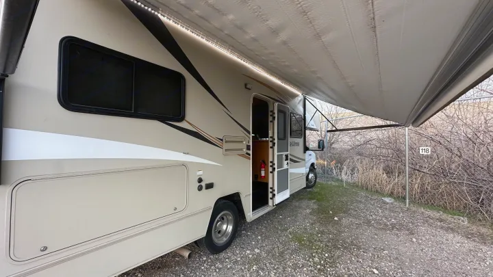 Beige Winnebago rv rental with extended awning & LED lights, parked on gravel beside dry bushes. Ideal for camping.. Winnebago Minnie Winnie 2018
