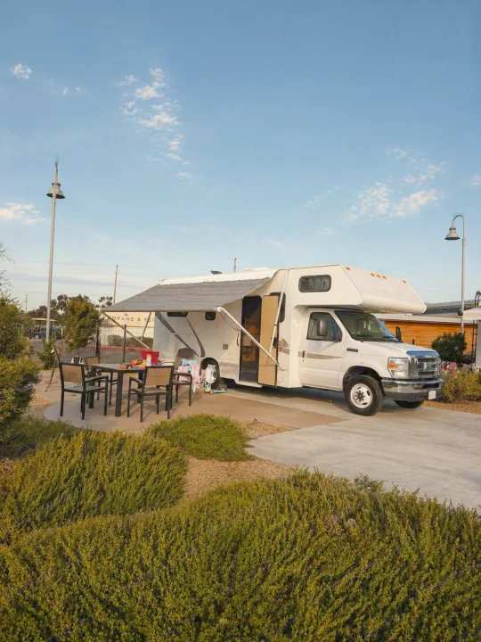 RV rental parked at a campsite with an extended awning and outdoor dining set, surrounded by green bushes under a blue sky.. Thor Majestic 2013