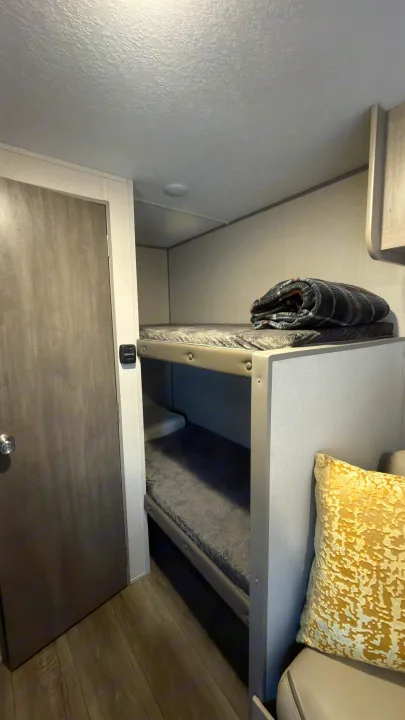 An rv rental interior with stacked bunk beds, a grey door, small thermostat, and a yellow cushion.. Coachmen Catalina Summit Series 7 16bhx 2024