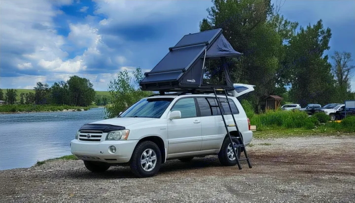 A white SUV with an open rooftop tent is parked by a scenic river, ready for a camping rv rental adventure.. Toyota Highlander 2005