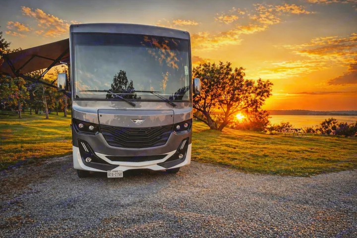 A sleek RV rental is parked on a gravel lot, facing a stunning sunset over a lake. Embrace nature & adventure on your next trip.. Fleetwood Fortis 2021