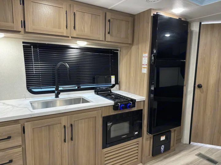 Inside an RV rental: a compact kitchen with a black faucet, stainless sink, stovetop, microwave, fridge, and light wood cabinets.. Keystone RV Coleman Light 2025