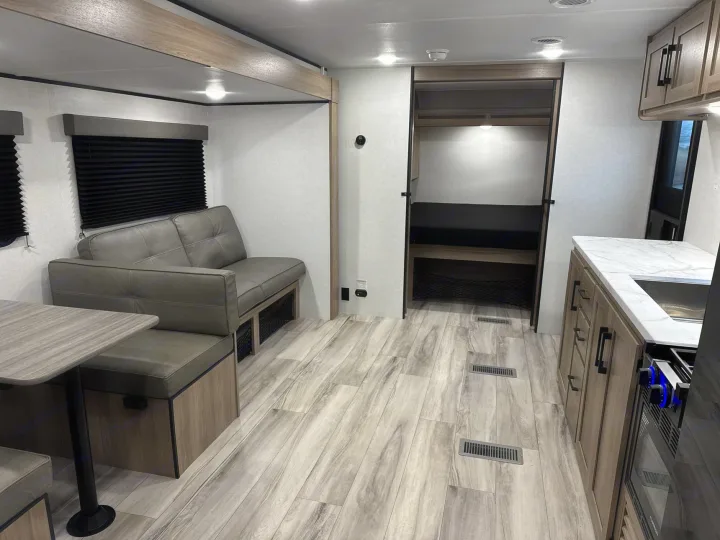 Modern rv rental interior: dinette, grey sofa, kitchenette with oven, and a bunk bed nook. Light wood floor.. Keystone RV Coleman Light 2025
