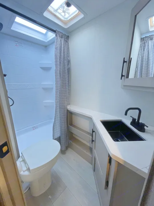 Compact RV rental bathroom with a white shower, toilet, and vanity featuring a black sink. Includes a patterned curtain & skylight.. KZ Connect 241BHKSE 2026