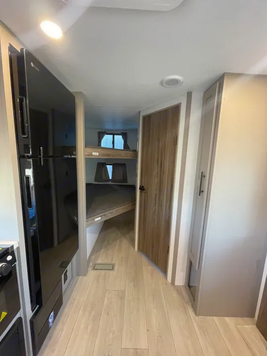 RV rental interior: A bright, modern hallway with light wood-look flooring leads past a black refrigerator to built-in bunk beds.. KZ Connect 241BHKSE 2026