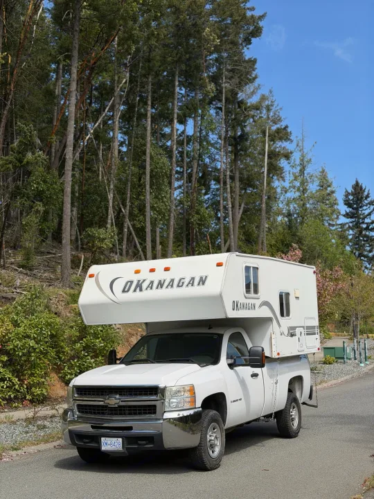 An Okanagan RV rental truck camper on a white Chevy Silverado, parked on a scenic road surrounded by tall trees and blue sky.. Chevrolet Silverado 2500HD 2008