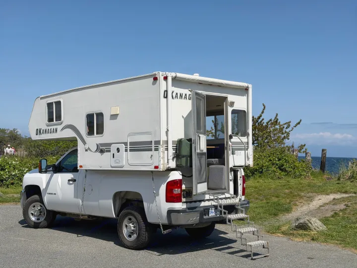 Sunny day with a white pickup truck, an Okanagan truck camper (potential rv rental) parked by the ocean. Open door and stairs visible.. Chevrolet Silverado 2500HD 2008