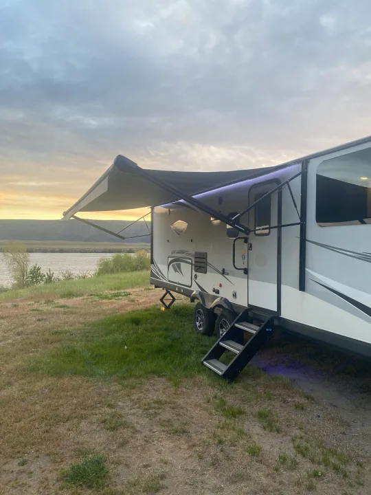 RV rental parked by a river at golden hour, with an illuminated awning and steps. Ideal for a scenic camping trip.. Jayco White Hawk 2020