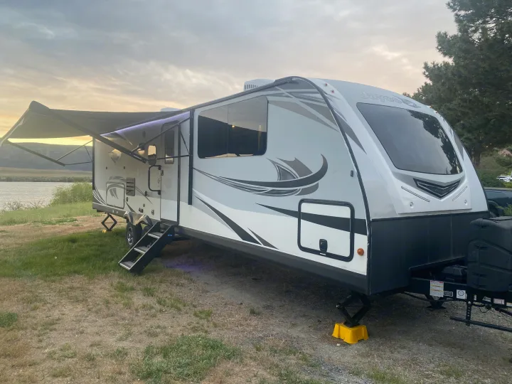 This RV rental offers a perfect lakeside camping spot at dusk. The white travel trailer has its awning extended and steps down.. Jayco White Hawk 2020