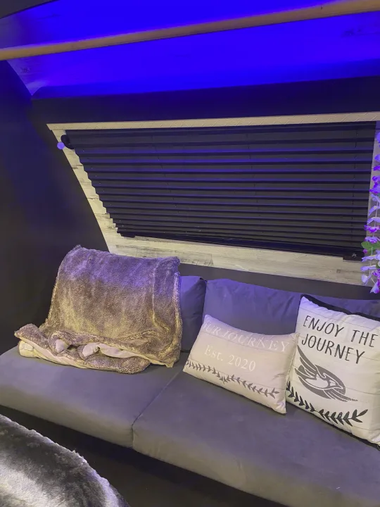 Cozy rv rental seating area featuring a grey couch, plush blanket, "Journey" pillows, window blinds, and ambient blue light.. Jayco White Hawk 2020