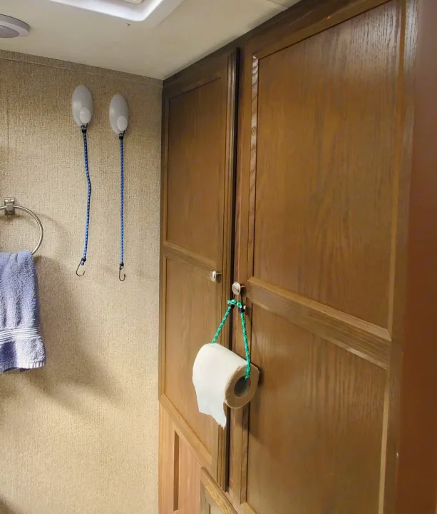 An RV rental bathroom showing a toilet paper roll hung from cabinet knobs with a bungee cord, near a vent fan and toilet.. Coachmen Apex Nano 2018