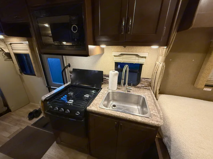 Compact RV rental kitchen featuring a microwave, stovetop, sink, and a cozy sleeping area, ideal for travel.. Thor Freedom Elite 2017