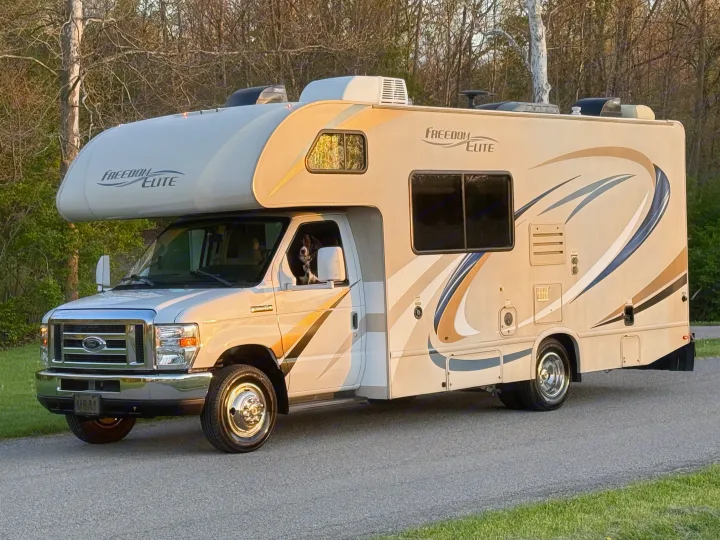 A Class C RV rental, a Freedom Elite model, is parked on a road with a dog looking out the driver's window. Trees line the background.. Thor Freedom Elite 2017