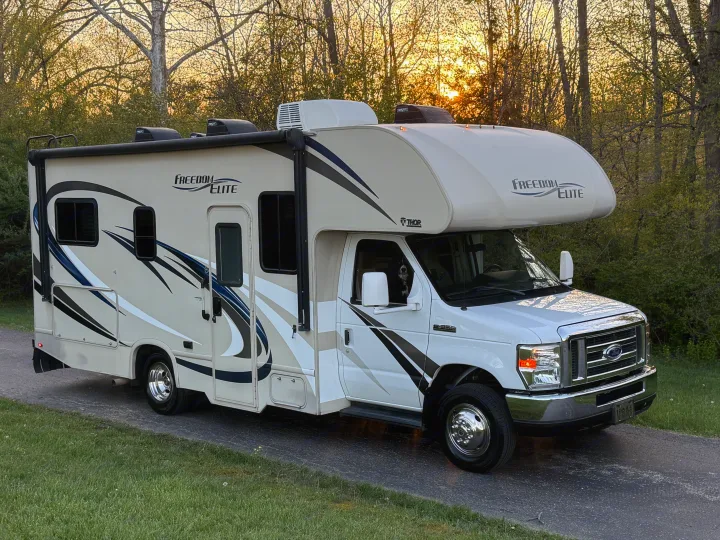 A white and tan Freedom Elite Class C RV rental, with a dog visible in the driver's window, is parked on a driveway at sunset.. Thor Freedom Elite 2017