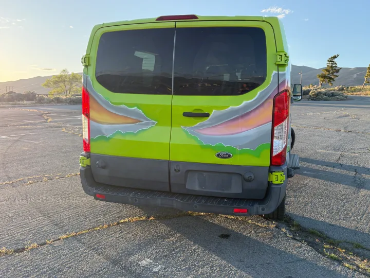 Bright green custom Ford Transit van, likely an RV rental, with a torn-look paint job, parked on asphalt at sunset with hills behind.. Ford Transit 2017