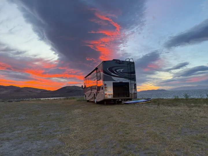An RV rental parked by a lake, with a paddleboard and mountains, under a breathtaking orange and red sunset.. Thor Palazzo 2014