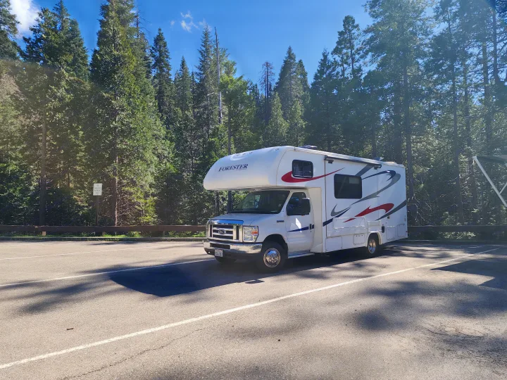 A white Forester RV rental is parked in a wooded parking spot, surrounded by tall evergreen trees under a clear blue sky.. Forest River Forester 2018