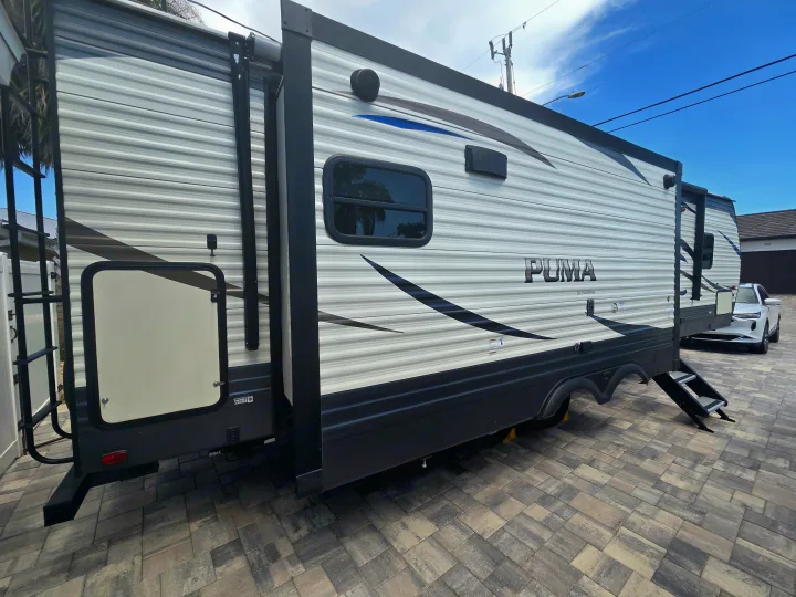 Side view of a PUMA travel trailer RV rental on a paved drive, with its slide-out extended, steps down, and rear ladder.. Forest River Palomino Puma 2020