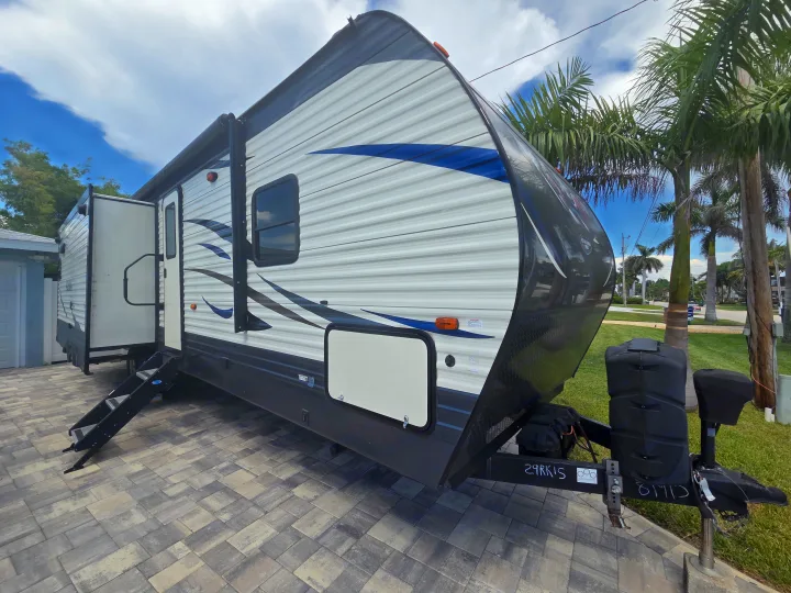White and blue travel trailer RV rental, parked on a paved driveway with steps extended and a slide-out visible. Palm trees in background.. Forest River Palomino Puma 2020