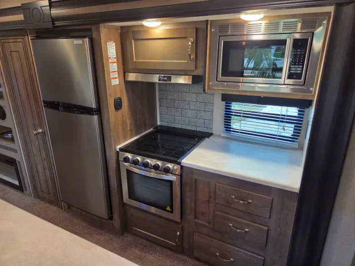 RV rental kitchen: stainless steel fridge, oven, microwave, gray backsplash, wood cabinets, and window with blinds.. Forest River Palomino Puma 2020